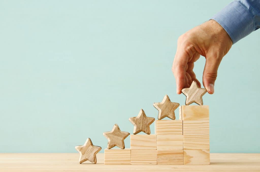 light wooden blocks stacked next to each other, elevating in height. Wood block star on each level, indicating growth with a hand placing the fifth and tallest star on light blue background.